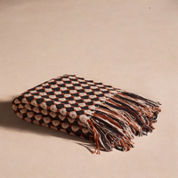 The Hampton Cotton Throw by Meso Goods is a handloom flatweave in black, beige, and brown with a textured honeycomb pattern and long wool fringe on one end, shown folded on a neutral surface.