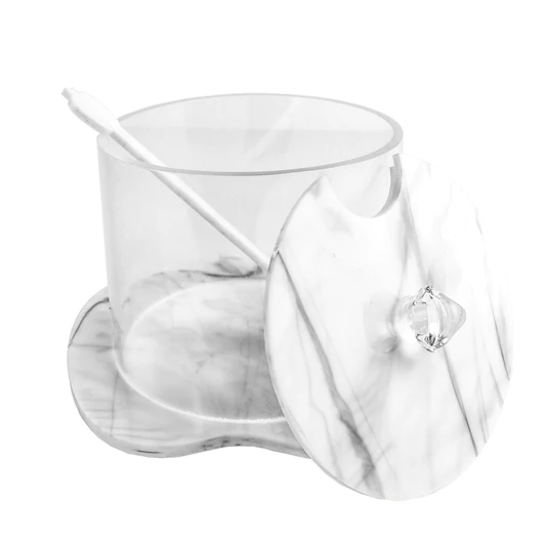 The BT- Shalom Lucite Apple Honey Jar features a clear cylindrical design with marble accents on the white lid and base. The slightly open lid reveals the jar's interior, and it comes with a spoon for serving honey.