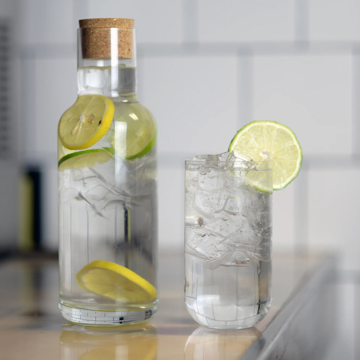 A Luigi Bormioli Sublime DOF tumbler from the set of 4, filled with ice water and garnished with lemon and lime slices, sits on a countertop before a white tiled wall; all pieces are dishwasher safe for easy cleanup.