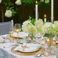 An elegant outdoor table features Kim Seybert’s Luminance Napkin in White & Gold, white and gold plates, gold-rimmed glasses, white florals, tall white candles, and lush greenery for a sophisticated setting.