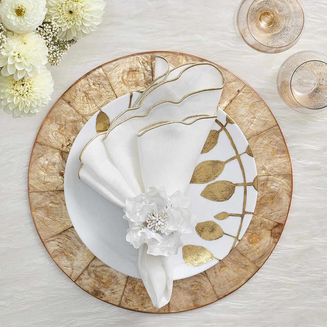 A decorative table setting features the Kim Seybert Luminance Napkin in White & Gold, folded in a floral napkin ring on a gold-edged plate and charger, with champagne glasses and white flowers nearby. Set includes 4 napkins.
