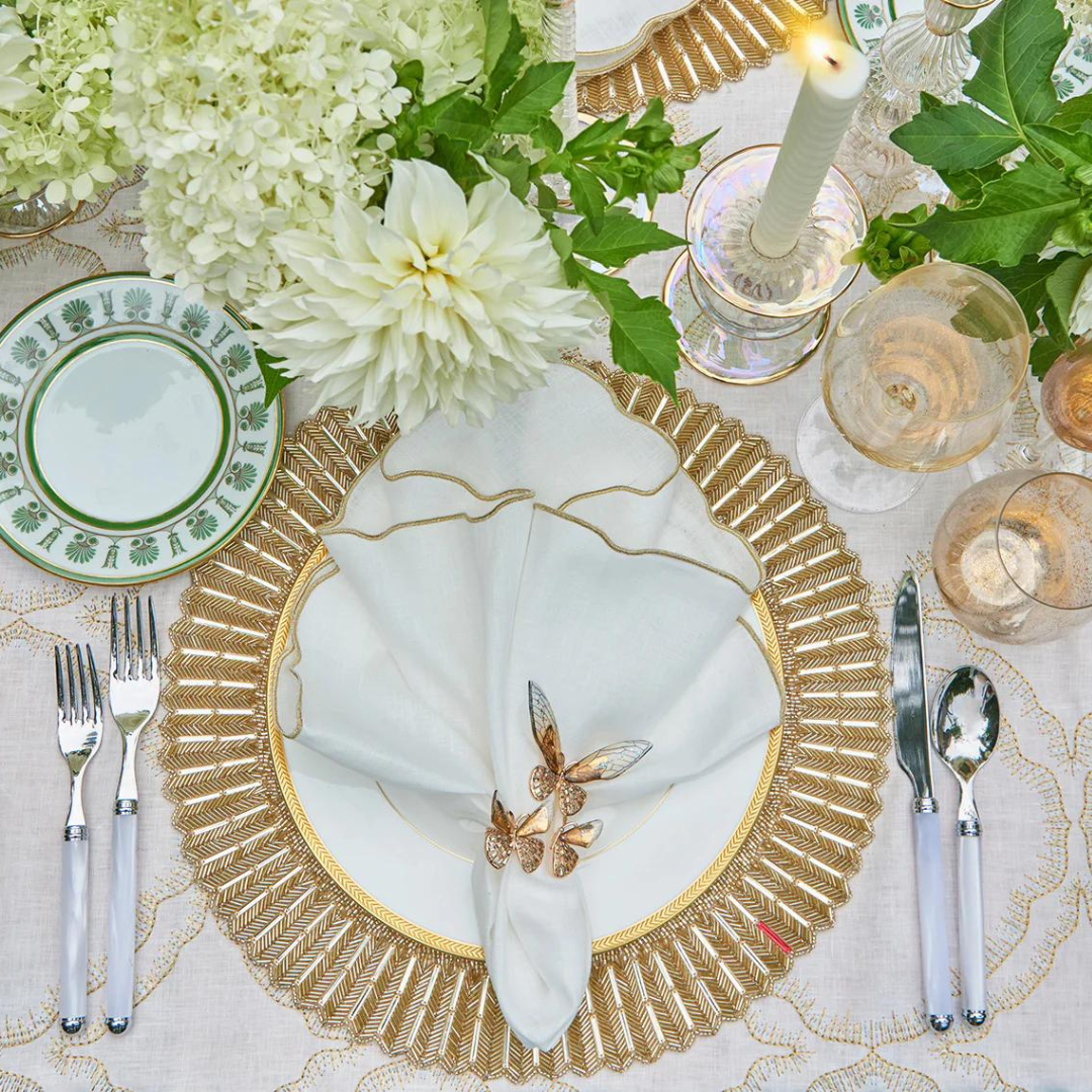Elegant table setting featuring Kim Seybert's Luminance Napkin in White & Gold, paired with a gold placemat, white and gold plate, floral napkin ring, cutlery, glassware, green-accented china, white flowers, and a lit candle.