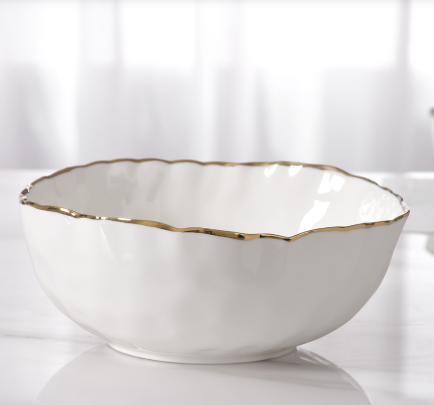 The PAMPA BAY Portofino Serving Bowl, crafted from white porcelain with a scalloped gold trim, is part of the elegant oven to table collection and sits beautifully against a soft, blurred background.