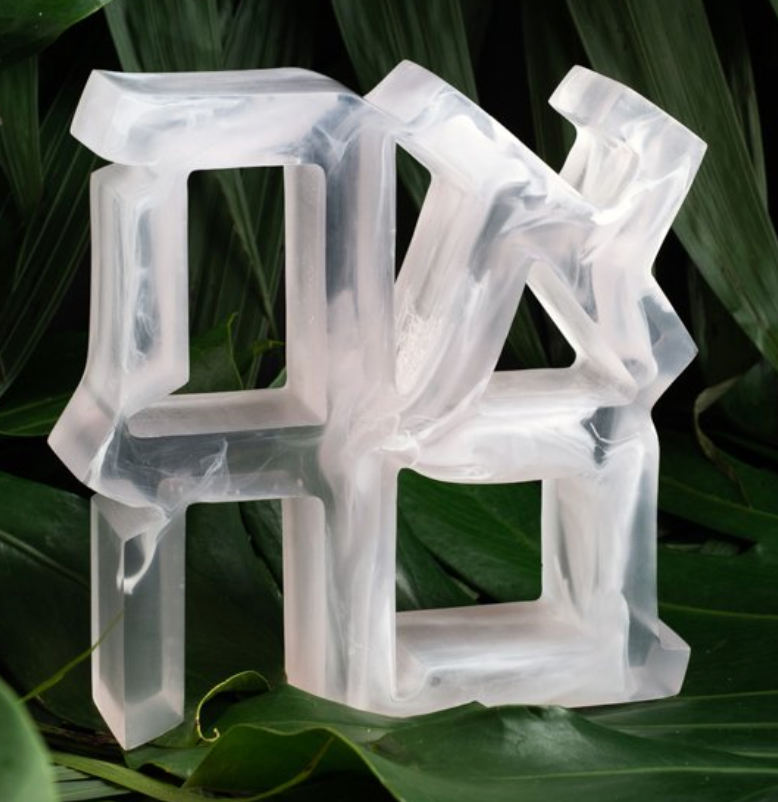 A translucent resin Ahava Sculpture by CDMX / BRUCI, with geometric shapes and curved edges, sits on large green leaves. The piece features a frosted, marble-like texture. BACK IN STOCK.
