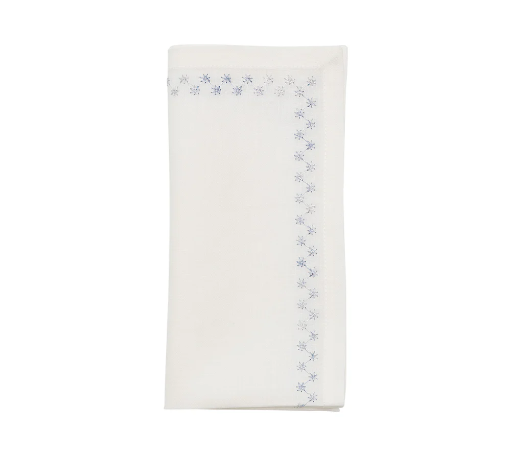 The Kim Seybert Starry Night Napkin Set of 4 in White & Blue features neatly folded white linen napkins with blue embroidered snowflake borders and delicate metallic embroidery on two edges, displayed on a plain white background.