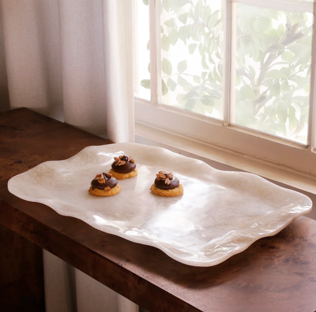 The Beatriz Ball Resin Tray Alabaster Extra Large displays three decorated cookies on a wooden surface near a window, with sheer curtains and leafy greenery visible outside.