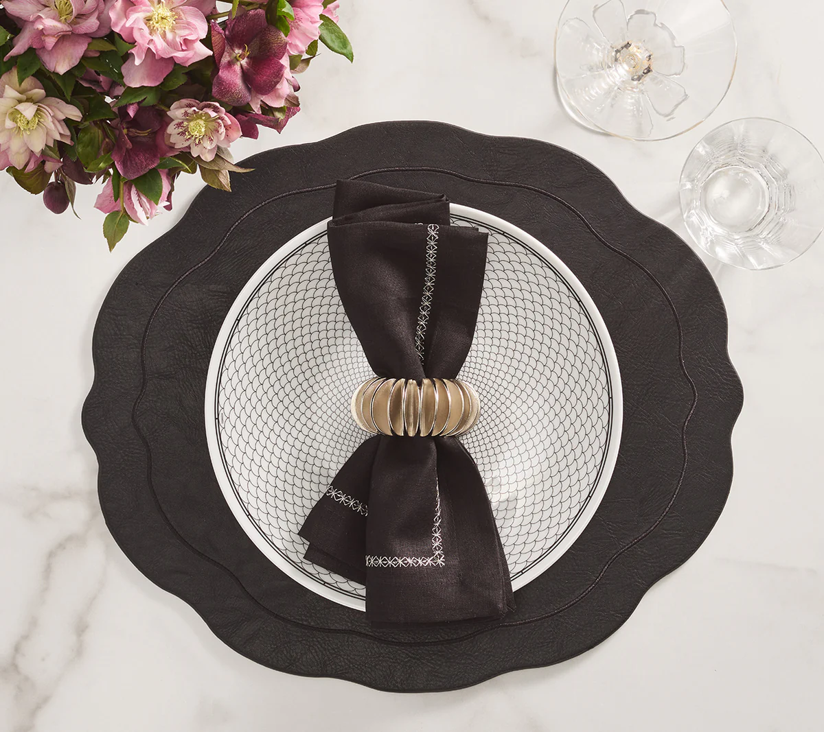A black scalloped placemat holds a white patterned dinner plate and black napkin, accented with the Kim Seybert Demilune Napkin Ring Silver Set of 4, on marble beside a glass and purple flowers for a sculptural table setting.