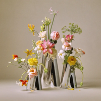 Five Georg Jensen Lituus Vases, each hand-polished from stainless steel and crafted in varying heights, display an artistic arrangement of colorful lilies, roses, and daisies against a neutral background. Scattered petals and leaves add a touch of natural elegance to the scene.