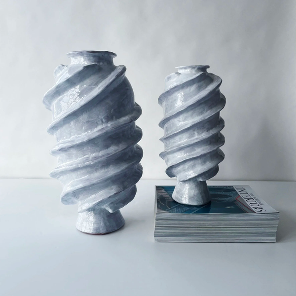 Two spiral-patterned gray ceramic vases of different heights sit on a white surface; one rests on the table, the other atop "Interiors" magazines, both reflecting the Ithaka Vase by Ellen Robinson Studio.