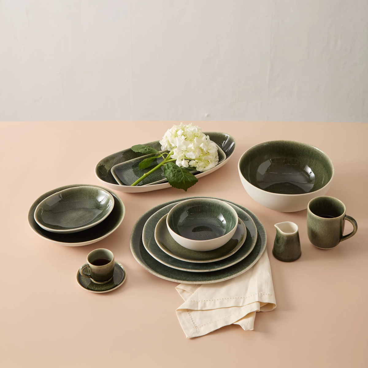 The Maguelone Orage Dinnerware by Jars - Rosenthal features handcrafted dark green stoneware dishes and cups, beautifully arranged on a peach surface and accented with white hydrangeas.