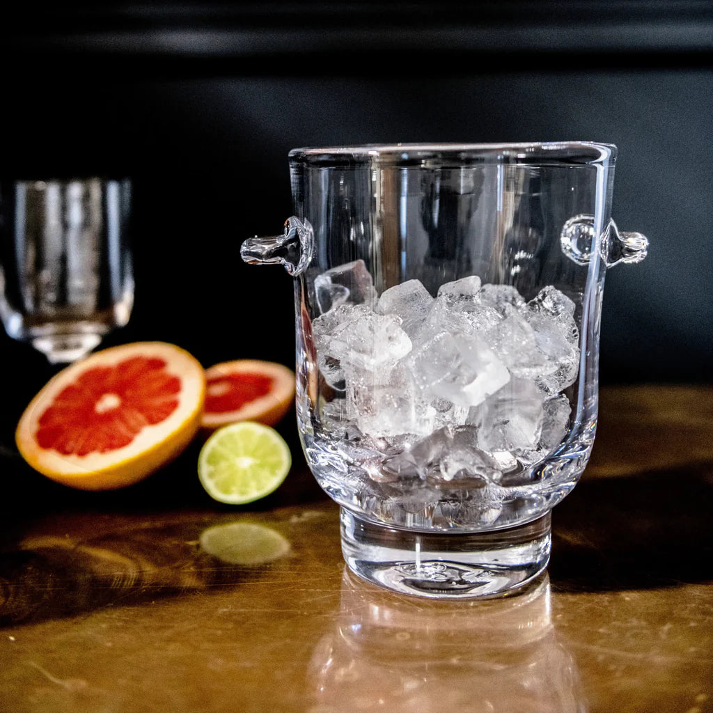 The Simon Pearce Manchester Ice Bucket, filled with ice cubes, rests on a reflective surface; behind it are a halved grapefruit, lime, and a blurred wine glass against a dark background.