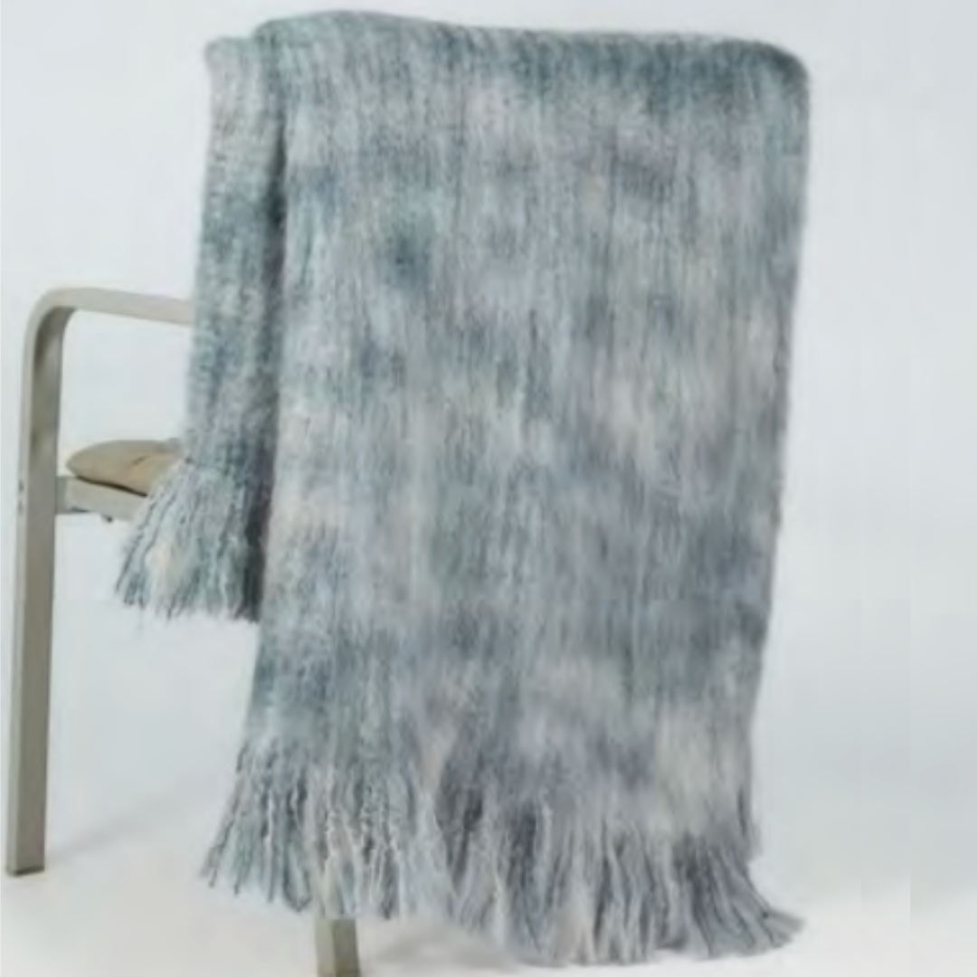 The Mantas Ezcaray 5th Elements Throw—a soft, blue-gray fringed blanket made with heirloom-quality textiles—is draped over a light chair, its long tassels cascading toward the floor.