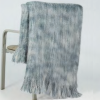 The Mantas Ezcaray 5th Elements Throw—a soft, blue-gray fringed blanket made with heirloom-quality textiles—is draped over a light chair, its long tassels cascading toward the floor.