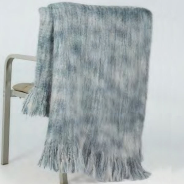 The Mantas Ezcaray 5th Elements Throw—a soft, blue-gray fringed blanket made with heirloom-quality textiles—is draped over a light chair, its long tassels cascading toward the floor.