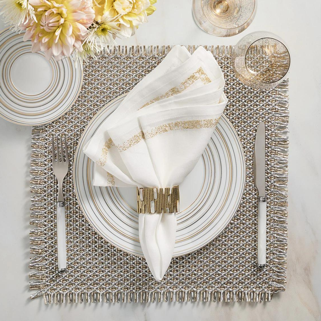 Elegant table setting featuring a white and gold-rimmed plate, folded white napkin adorned with the Matrix Napkin Ring in Gold by Kim Seybert, silverware, wine glass, fresh flowers, and a textured placemat.