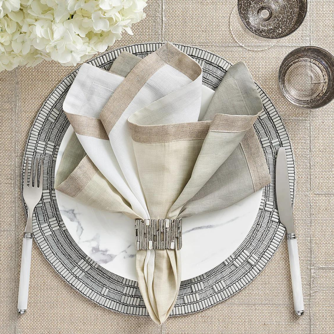 A place setting featuring a marble-patterned plate, silver cutlery, and a folded multicolored napkin elegantly held by the Kim Seybert Matrix Napkin Ring Set of 4 in Gold, arranged on a textured placemat. Glassware and white flowers complete the sophisticated display.