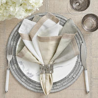 A place setting featuring a marble-patterned plate, silver cutlery, and a folded multicolored napkin elegantly held by the Kim Seybert Matrix Napkin Ring Set of 4 in Gold, arranged on a textured placemat. Glassware and white flowers complete the sophisticated display.