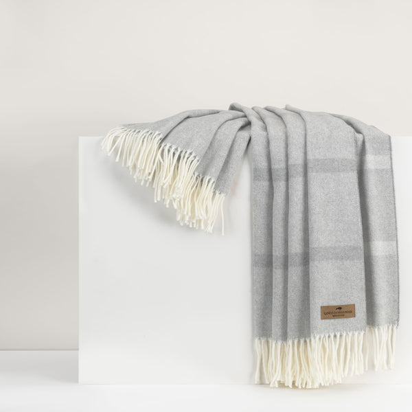 A light gray wool reversible throw with a plaid pattern and cream fringe, like the Montauk Stripe Throw by Lands Downunder, is draped over a white surface.