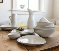 The Simon Pearce Barre Dinnerware Alabaster set—white ceramic dishes, bowls, cups, and a pitcher—is neatly arranged on a wooden dining table in a bright room with a window in the background.