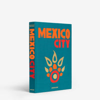 A teal book cover from Assouline’s Travel Series - Mexico City features bold orange text and a stylized orange-gold paw print evoking Spanish-baroque style. The spine also displays MEXICO CITY vertically, nodding to colonial architecture.