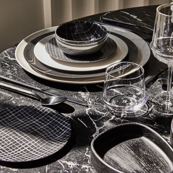 A modern table setting on black marble showcases Serax's Zuma Dinnerware Pacific—porcelain plates with Bauhaus-inspired grids, matching bowls, two clear glasses, and neatly arranged black cutlery.