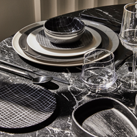 A modern table setting on black marble showcases Serax's Zuma Dinnerware Pacific—porcelain plates with Bauhaus-inspired grids, matching bowls, two clear glasses, and neatly arranged black cutlery.