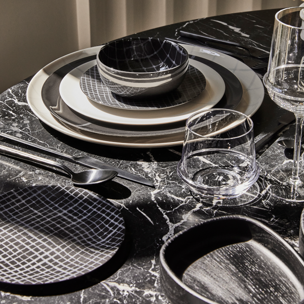 A modern table setting on black marble showcases Serax's Zuma Dinnerware Pacific—porcelain plates with Bauhaus-inspired grids, matching bowls, two clear glasses, and neatly arranged black cutlery.