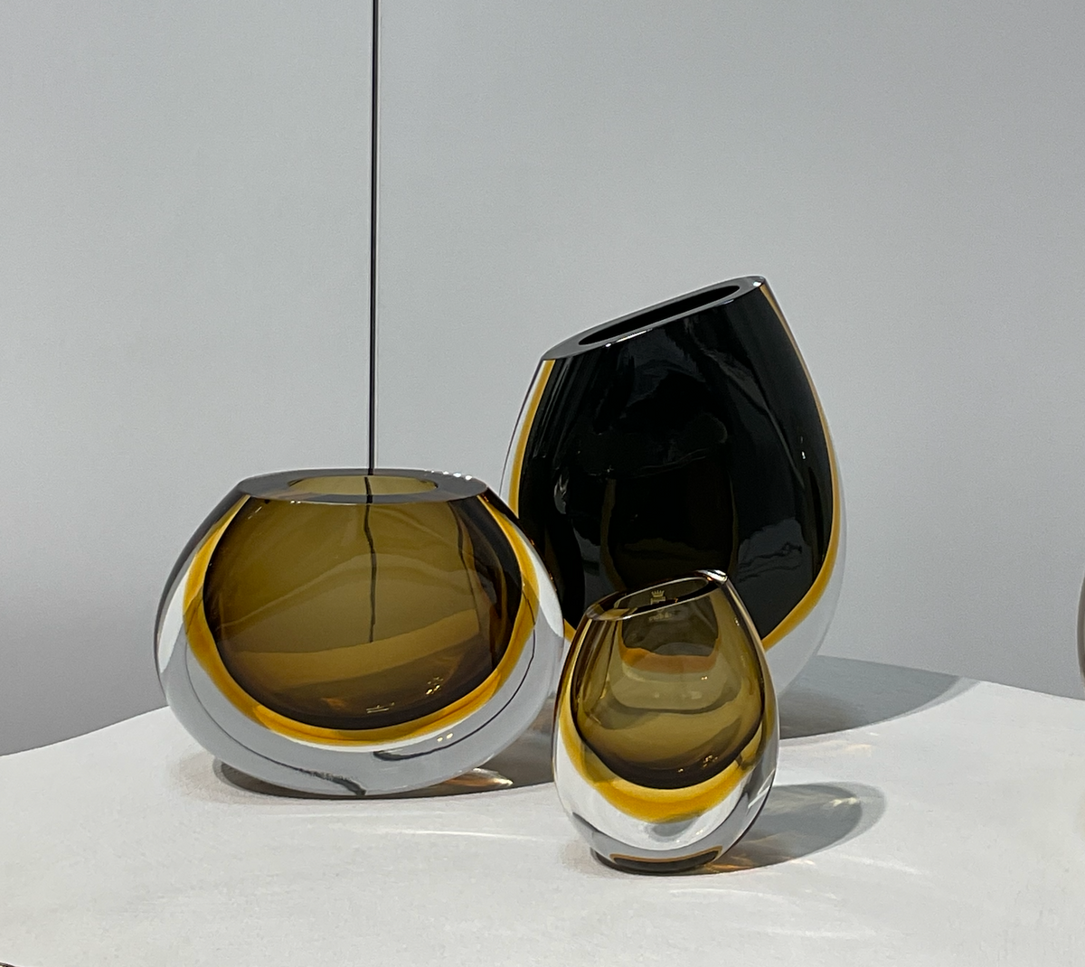The Gardeco Vase 96 - Black Amber features a modern, round design with organic, asymmetrical shape and polished finish. It displays clear, amber, and black tones, sitting on a white surface against a plain gray background.