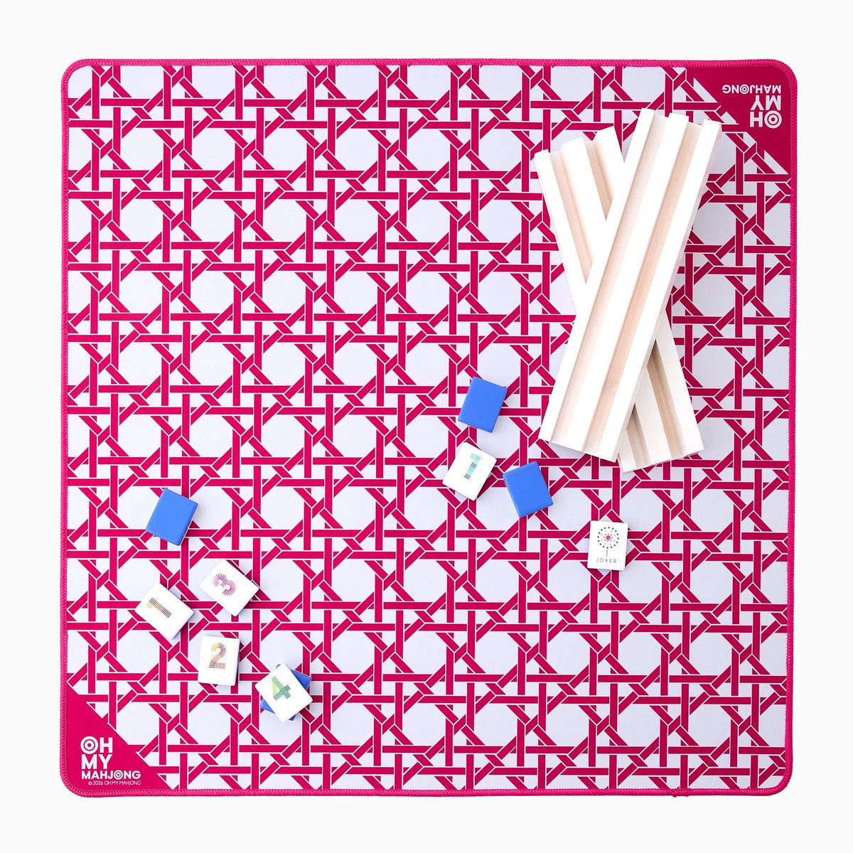 The Oh My Mahjong Travel Mini Mahjong Game Mat (22” x 22”) features a pink geometric pattern, white racks on the right, blue tile holders and tiles on the left, and “Oh My Mahjong” printed in the bottom left—ideal for portable play.