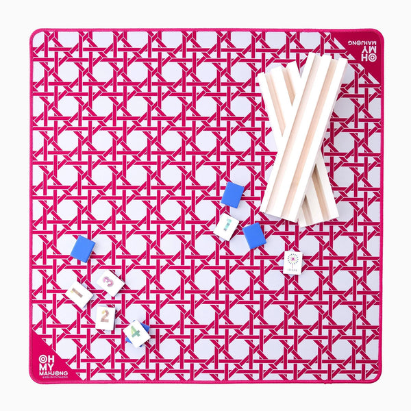 The Oh My Mahjong Travel Mini Mahjong Game Mat (22” x 22”) features a pink geometric pattern, white racks on the right, blue tile holders and tiles on the left, and “Oh My Mahjong” printed in the bottom left—ideal for portable play.