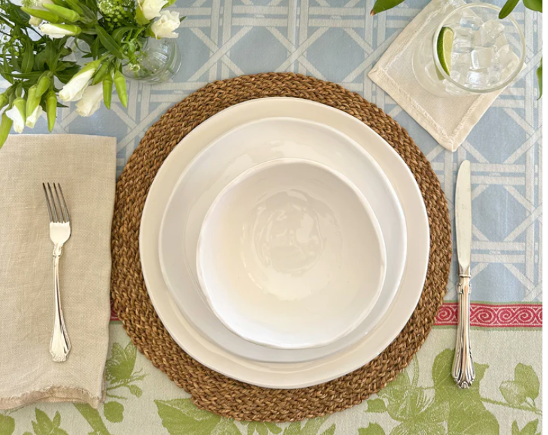 A table is set with a woven placemat, Relish Simple White Melamine Dinnerware (best seller, dishwasher safe), a linen napkin with fork left, knife and spoon right, a glass of water with lime, and white flowers on a patterned tablecloth.