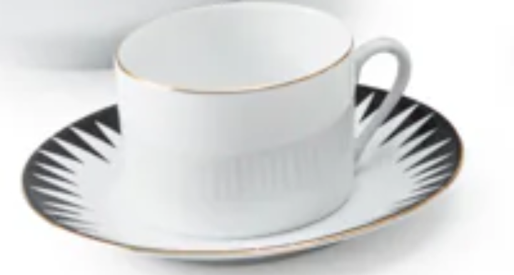 The Punk Dinnerware teacup by Molecot, crafted from white Limoges porcelain with a gold rim, rests on a matching saucer adorned with a black and white geometric starburst edge for a modern, elegant table setting.