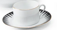 The Punk Dinnerware teacup by Molecot, crafted from white Limoges porcelain with a gold rim, rests on a matching saucer adorned with a black and white geometric starburst edge for a modern, elegant table setting.