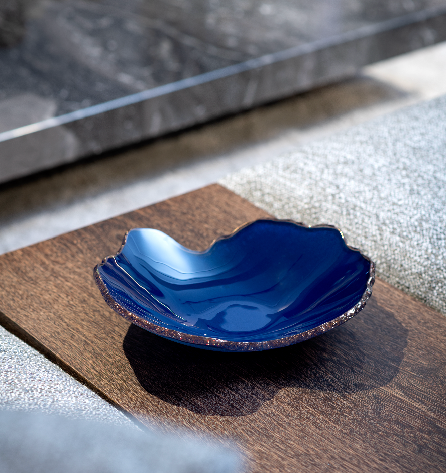 A deep blue, irregularly shaped Mapa Azul With Bronze Trim Bowl by Gardeco sits on a wooden surface with textured grey and stone accents in soft sunlight, its wavy edge and bronze trim making it striking decor.