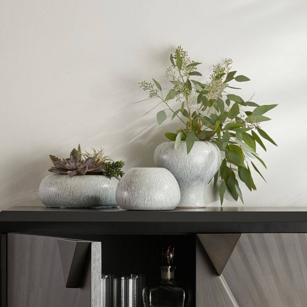 A trio of Dandy Vases s/3 - Grey by Arteriors, featuring an icy morn finish, are arranged on a modern, geometric cabinet with partially open doors. Two of the light gray ceramic vases display green plants—one filled with succulents and the other with leafy stems—evoking a spa-like sense of serenity.