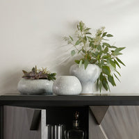A trio of Dandy Vases s/3 - Grey by Arteriors, featuring an icy morn finish, are arranged on a modern, geometric cabinet with partially open doors. Two of the light gray ceramic vases display green plants—one filled with succulents and the other with leafy stems—evoking a spa-like sense of serenity.