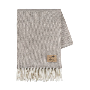 The Juno Cashmere Throw by Lands Downunder is a folded light grey luxury wool blanket with a subtle herringbone pattern, cream fringed edges, and a brown leather patch label in one corner.