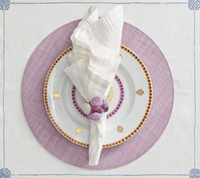 A white napkin in a Kim Seybert Sea Stone Napkin Ring Set of 4 - Lilac rests on a white plate with gold accents, atop a lavender charger and a white tablecloth with light blue embroidered corners.