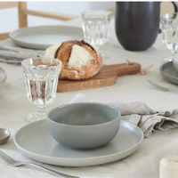 A neatly set dining table features Costa Nova's Pacifica Dinnerware in Oyster Grey, paired with a glass of water, cutlery, a linen napkin, and a wooden board with bread—creating a simple and inviting scene.