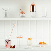 A minimalist bar setup with the Hepburn Glass Set of 4 by Nude Glassware in lead-free crystal, copper bar tools, assorted glassware, berry and orange cocktails, and a dish of small orange fruits against a white tiled wall.