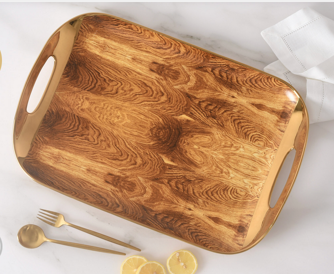 The PAMPA BAY Madera Gold Handle Tray, featuring a rustic wood design and cutout handles, rests on a marble surface beside a folded white napkin, gold cutlery, and sliced lemon.