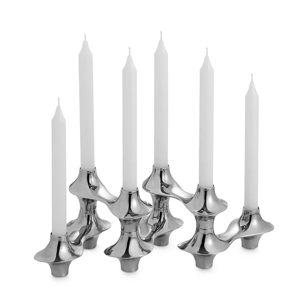Six white candles sit in modern, silver-colored holders inspired by Nambe's Cortina Hinged Candelabra, featuring a sculptural, modular design reminiscent of an adjustable candelabra on a white background.