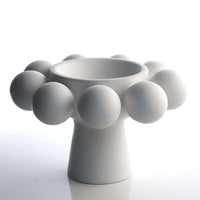The KleinReid Nimbus Footed Bowl Alabaster is a modern white ceramic piece with a flared base, round bowl, and large hand-appliquéd porcelain spheres spaced around the rim, shown on a reflective white surface.