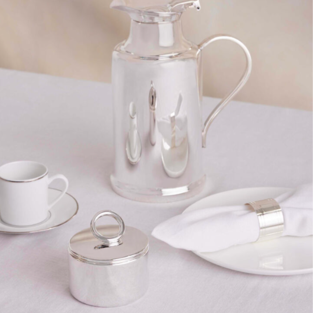 A Christofle Vertigo Silver Plated Sugar Bowl with handle, a silver pitcher, a white cup and saucer, and a white plate with napkin in a silver ring are arranged on a white tablecloth.