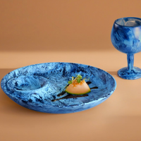 The Nashi Home Swirl Colored Resin Chip & Dip, a blue marbled platter, holds melon with herbs and sauce beside a matching goblet on beige—ideal for stylishly serving guests.