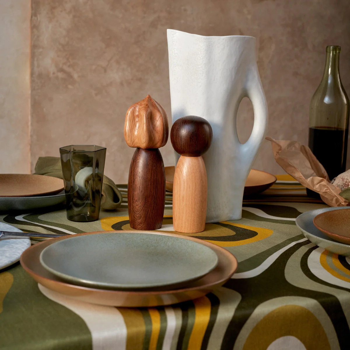 A dining table set with ceramic plates, a green glass, a wine bottle, a loaf of bread, L'Objet Picanto Salt + Pepper Mills (set of 2), a white sculptural vase, and wooden figurines on a bold retro geometric tablecloth.