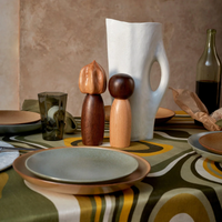 A dining table set with ceramic plates, a green glass, a wine bottle, a loaf of bread, L'Objet Picanto Salt + Pepper Mills (set of 2), a white sculptural vase, and wooden figurines on a bold retro geometric tablecloth.