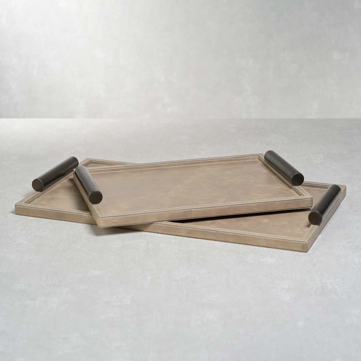 Two rectangular beige trays with raised black cylindrical handles are stacked at an angle on a light grey surface. The Zodax Belle Rectangle Leather Tray set, crafted in luxurious mink leather, seamlessly blends elegance and functionality for all your entertaining needs.