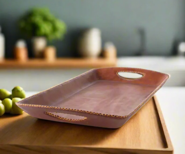 The Embroided Long Bar Tray - Brown by Bati Goods, featuring gold-trimmed edges and handles, sits atop a wooden surface. Crafted from organic tanned leather for a luxurious touch, its water-resistant base is adorned with green grapes, while a blurred kitchen background completes the scene.