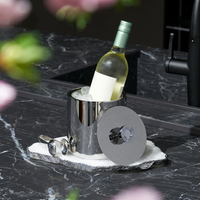 A bottle of white wine chills in the Striations Silver Ice Bucket with Tongs by Anna New York, its double-walled stainless steel design and matching lid resting on a marble slab, with blurred pink flowers framing the scene.